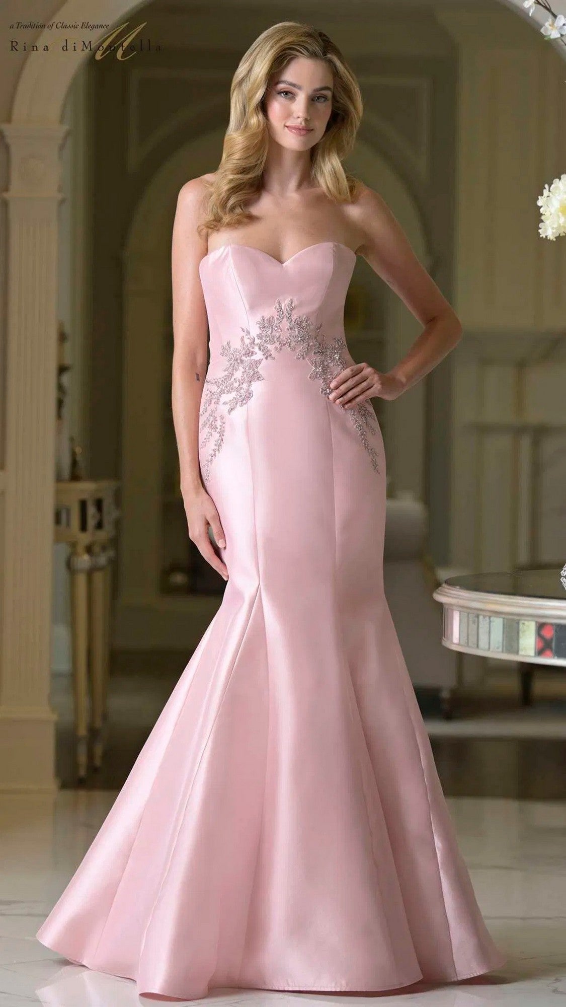 A woman stands indoors wearing the Rina diMontella RD3420, a long formal strapless fitted gown in light pink with silver floral embroidery and a flared hem.